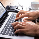 Person's Hand Typing On Laptop Over Wooden Desk